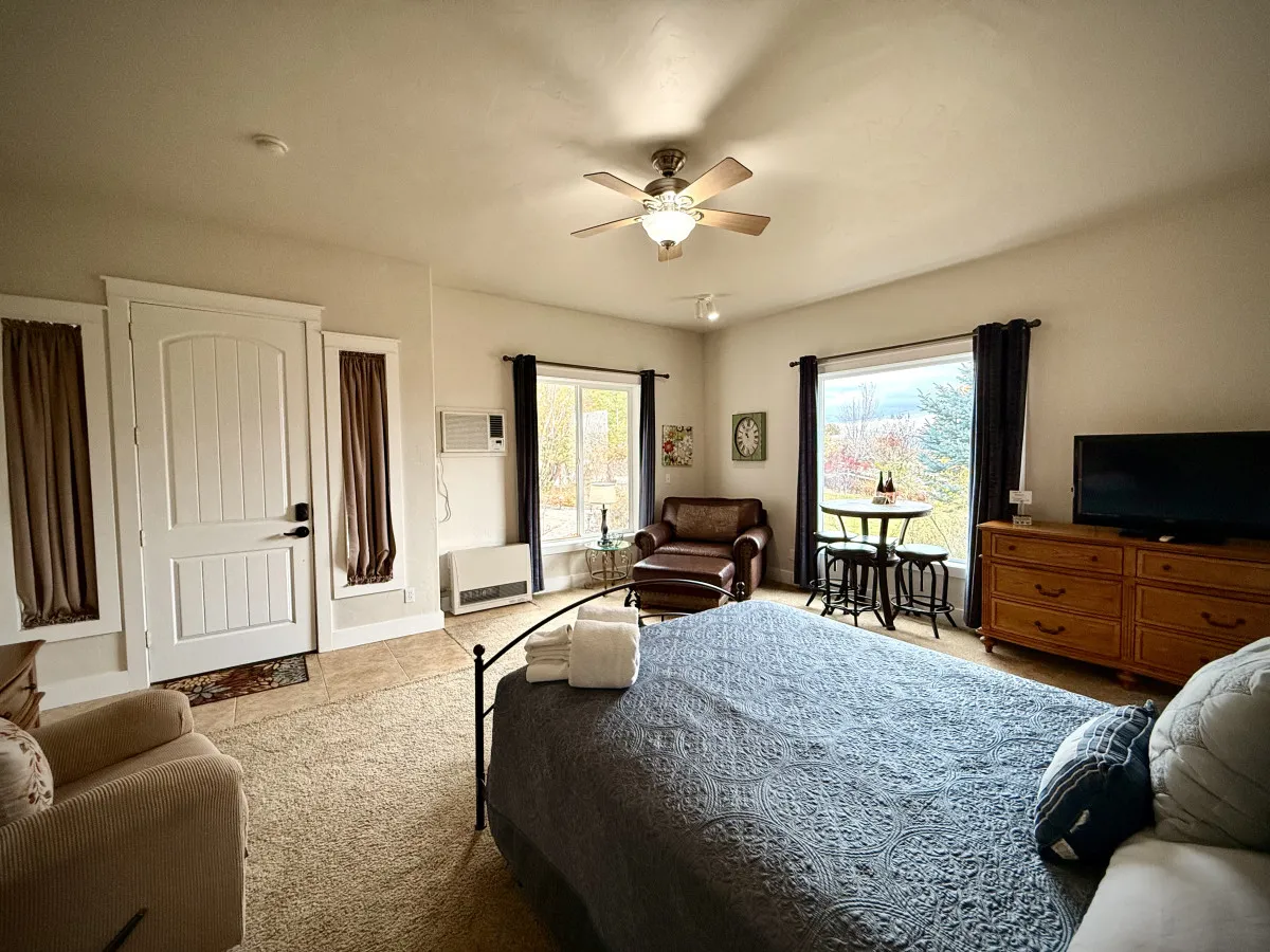 This image shows a bright, tranquil studio with a plush bed layered with decorative throw pillows, a comfortable armchair positioned for relaxation, windows that let in ample natural light and offer scenic views, and a small dining table ideal for intimate meals or morning coffee.