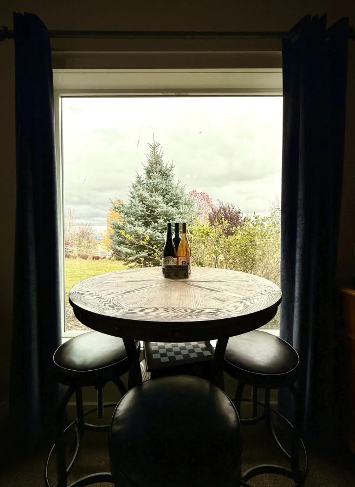 An inviting dining space with a round wooden table set for wine tasting or board games, surrounded by large windows with views of outdoor greenery. The warm lighting and welcoming layout make it perfect for shared meals or festive group celebrations.
