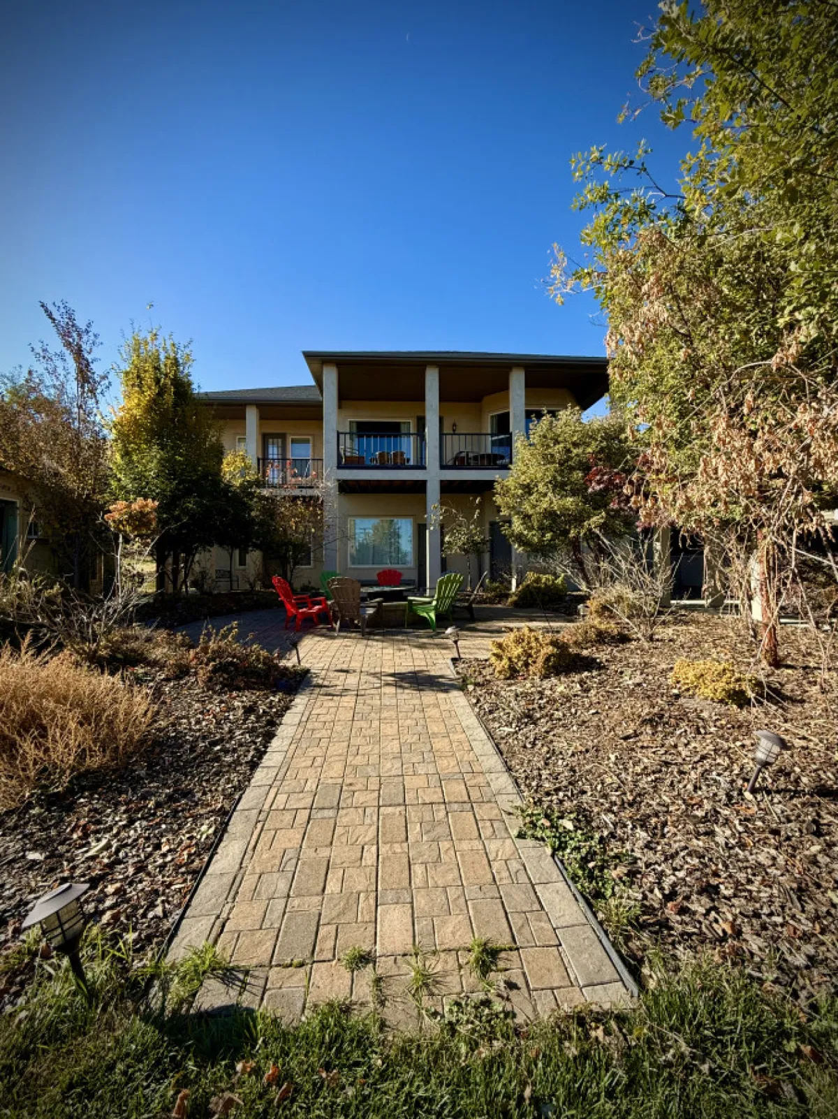 Two colorful chairs (one red, one green) are set on a patio near a pathway leading to the house. A balcony with seating is visible above, surrounded by lush greenery and offering a cozy spot to relax outdoors, possibly near a firepit.