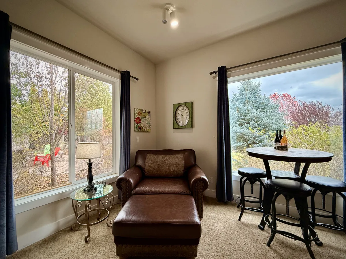 A tranquil interior living area featuring a leather chaise lounge, a small round table with wine bottles, and large windows overlooking lush greenery. The decor suggests a space perfect for relaxing with a book, socializing, or enjoying a glass of wine with friends.
