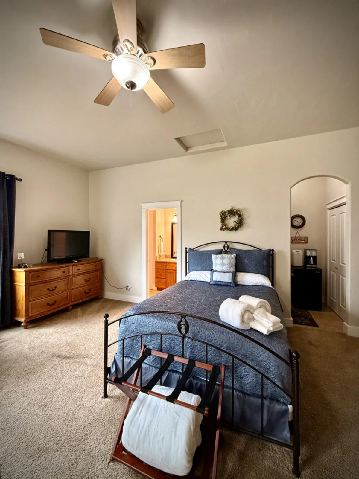 A relaxing bedroom setting featuring a bed with soft blue bedding, decorative pillows, a wooden dresser, TV, and neatly arranged towels. Soft morning light filters through curtains, creating a comfortable and inviting ambiance.