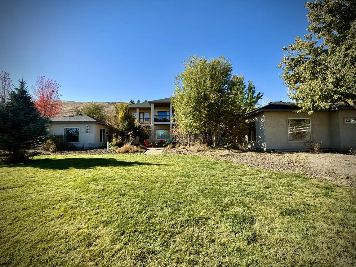A picturesque two-story house surrounded by a lush green lawn, mature trees, and a patio area with chairs set for outdoor gatherings. The peaceful setting is ideal for barbecues, sunset views, and evenings spent around a firepit.