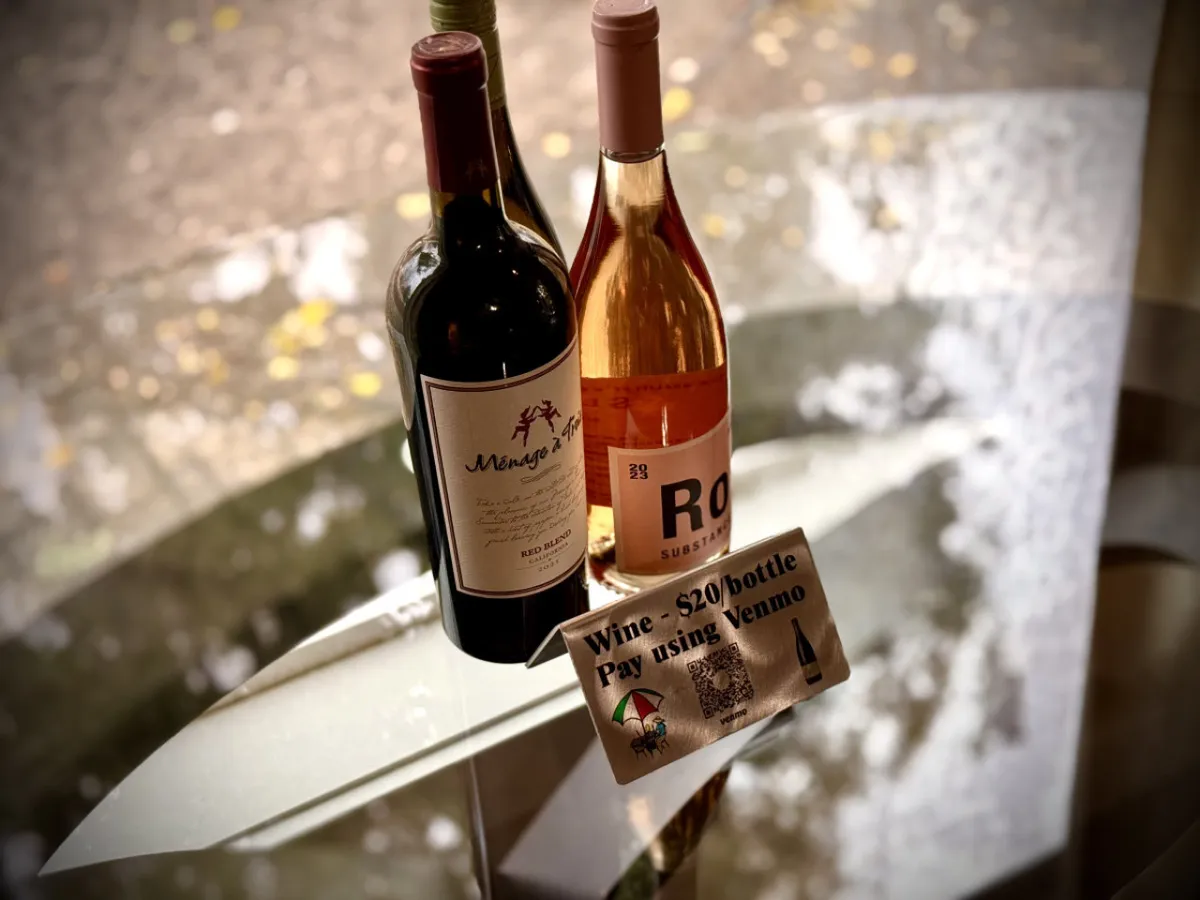 A cozy interior scene with two bottles of wine set beside a payment sign, suggesting social gatherings or wine tastings. The mood is casual and friendly, evoking the idea of sharing good times after a day of adventures.