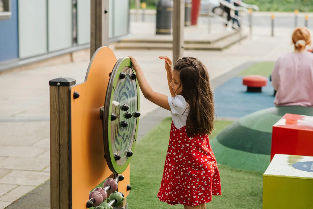 An outdoor playground scene featuring colorful equipment and a child joyfully playing. The area is vibrant and designed for interactive games, surrounded by greenery and open space, evoking a sense of youthful energy and family activity.