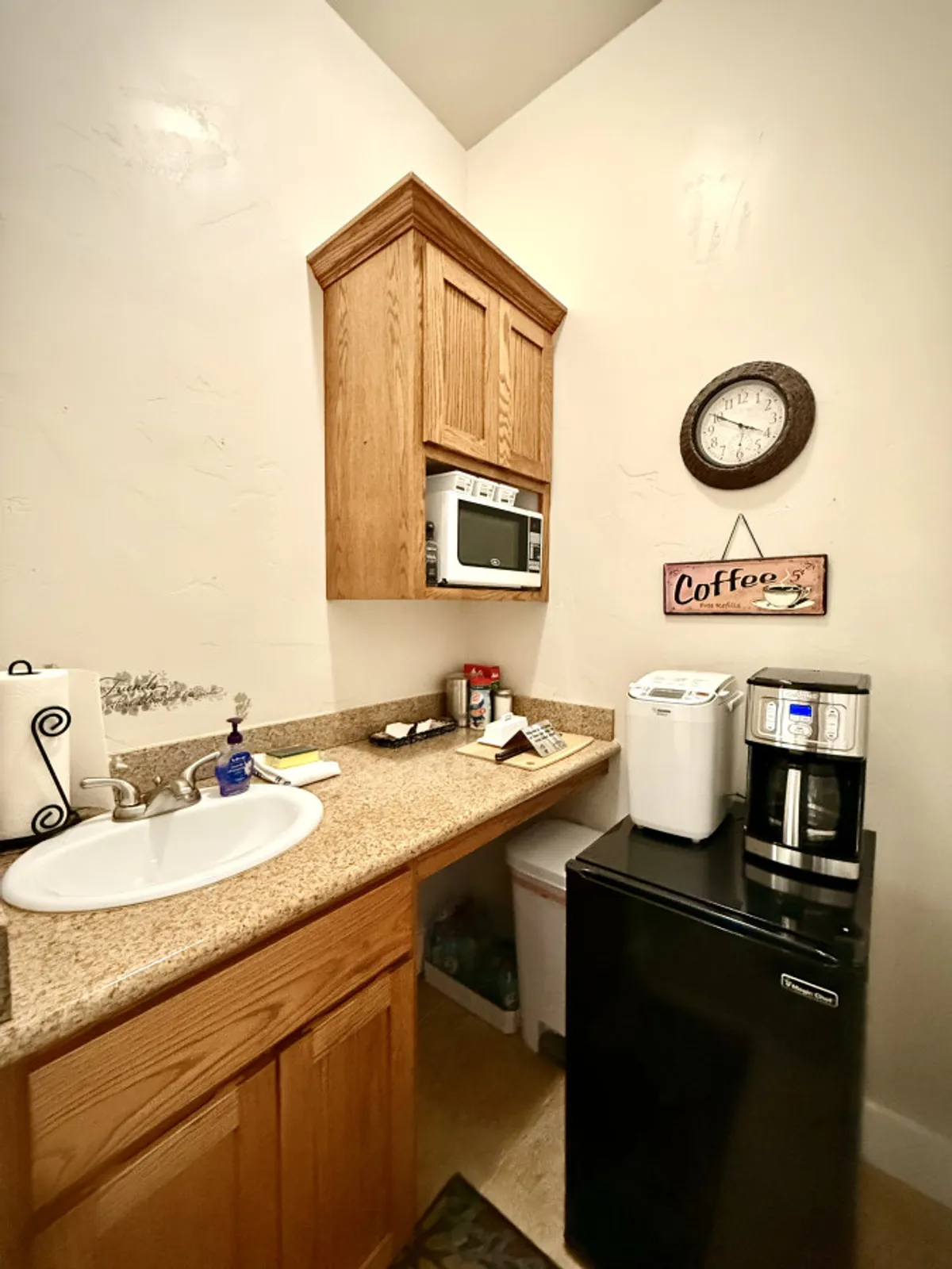 A compact, well-equipped kitchenette featuring a coffee maker, microwave, sink, wooden cabinets, and a wall clock. The space feels cozy and functional, perfect for brewing coffee or preparing light snacks at any time.