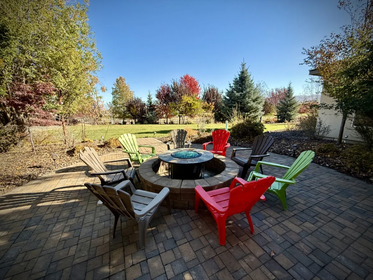 A cozy fire pit surrounded by colorful chairs in a beautifully landscaped yard, framed by lush greenery. The space is clearly designed for guests to gather, relax, and enjoy the outdoors together.