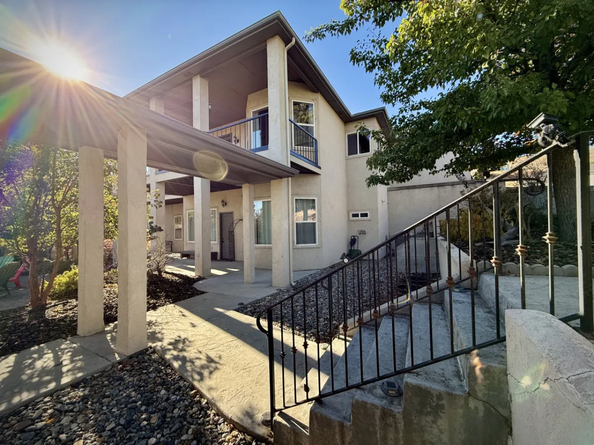 This exterior image features a two-story structure surrounded by lush greenery and sunlight filtering through the trees. The porch and garden area create a welcoming setting for outdoor gatherings, perfect for group activities and relaxation after a day of golf or skiing.