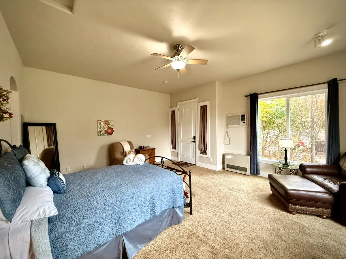 This inviting bedroom scene features a bed with blue bedding, a leather armchair beside a well-lit window with curtains, and a decorative painting on the wall. The atmosphere is calm and serene, with natural light streaming in, creating a sense of relaxation and comfort.